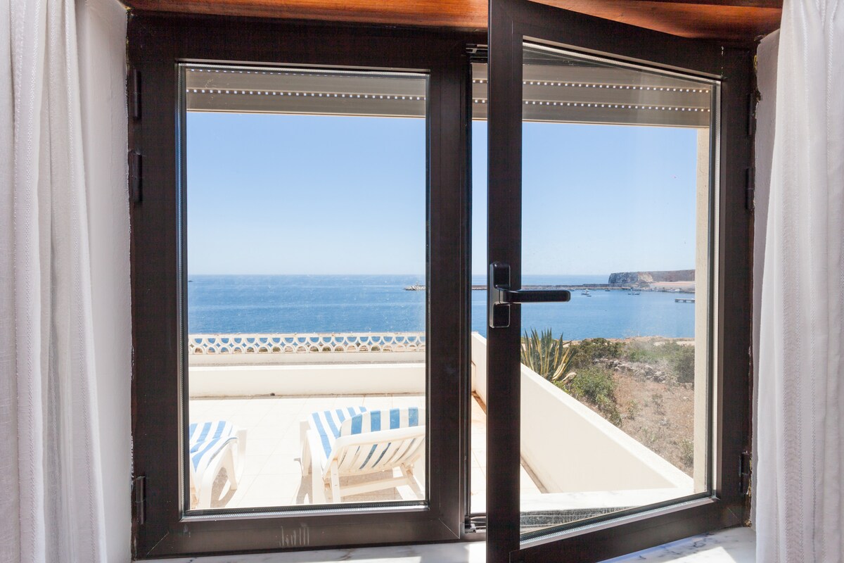 A large window offers a view of the ocean, with clear waters and a distant shoreline visible. Striped lounge chairs can be seen on the terrace outside, providing a relaxing spot to enjoy the scenery.