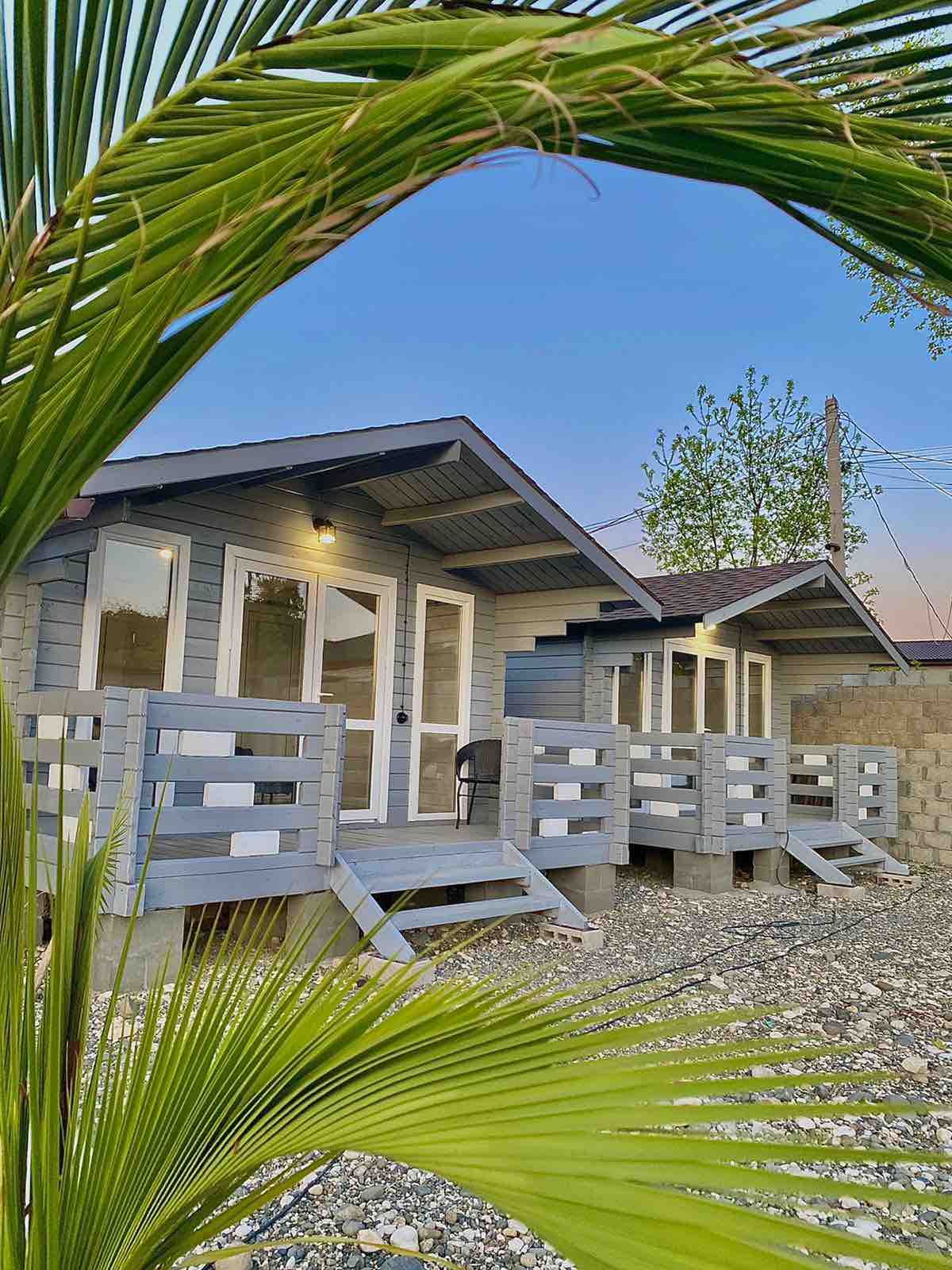 Two charming cabins are positioned under a clear sky, each featuring a front porch with steps. The natural scenery includes green palm fronds in the foreground, complementing the restful outdoor setting.