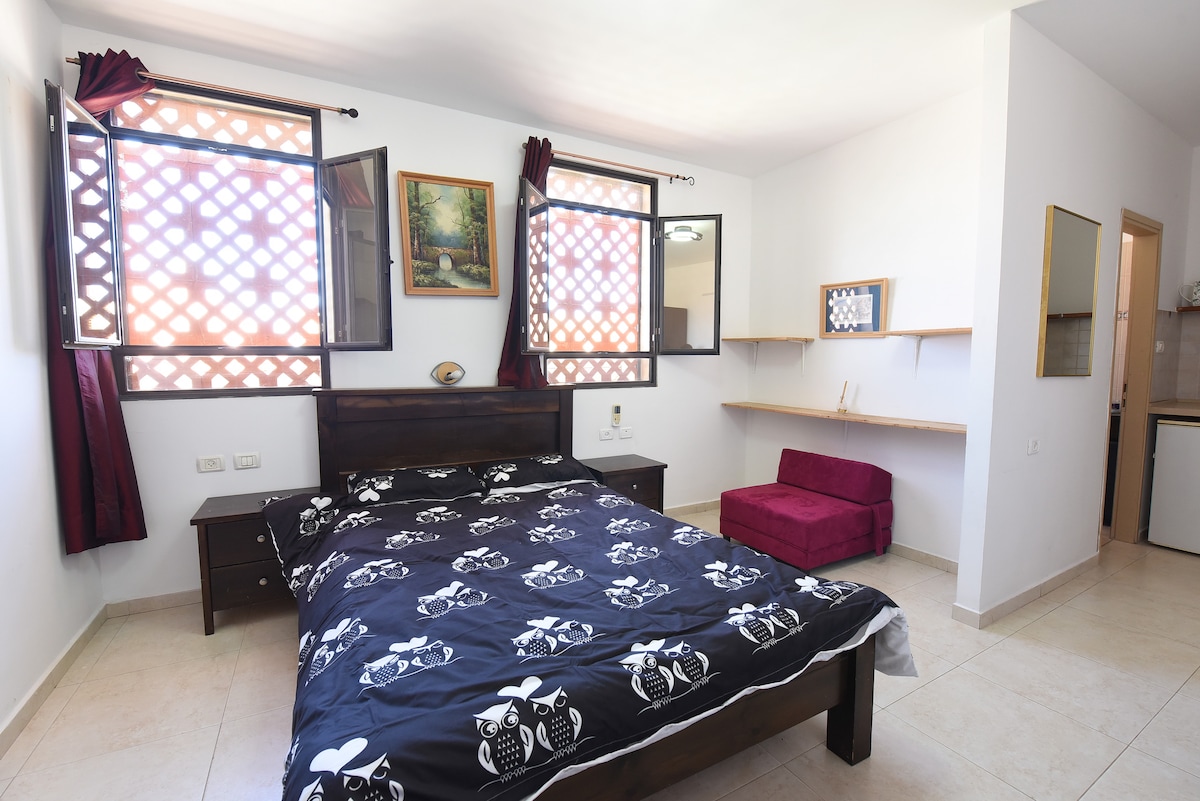 A double room features a spacious bed with a black and white patterned duvet. Natural light enters through two windows adorned with decorative grilles. A small seating area is positioned against the wall beside a wooden shelf. Wall art adds a touch of character to the space.
