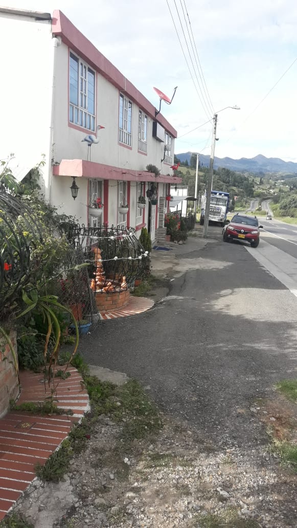 A two-story building is situated along a main road, featuring a bright exterior with decorative elements. A pathway lined with plants leads to the entrance. Vehicles can be seen on the road, with mountains visible in the background under a clear sky.