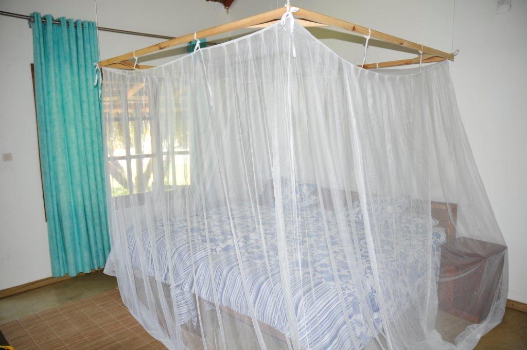 A spacious sleeping area features two beds draped with a mosquito net. Light-colored curtains frame a large window, allowing natural light to brighten the room. A wooden cupboard is visible, complemented by a light blue patterned bedspread.