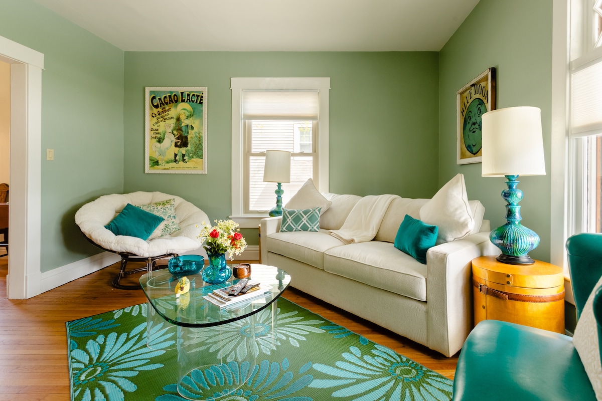 A light-filled living room features a large white sofa complemented by turquoise pillows. A round glass coffee table sits on a patterned area rug. Decorative wall art adds character, while a bright lamp illuminates the space. Natural light streams through partially covered windows.