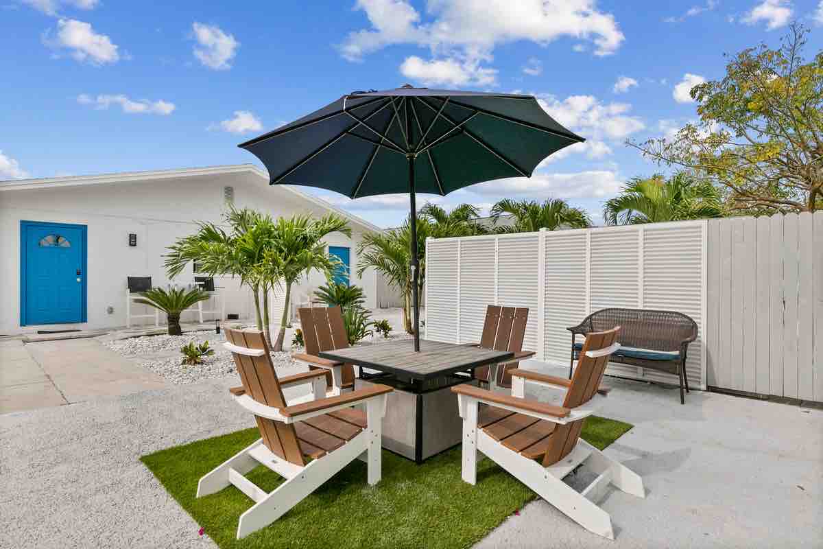 An outdoor seating area features a dark umbrella above a table surrounded by four wooden chairs. Lush greenery and white stones provide a serene backdrop, while a light-colored fence encloses the space, offering privacy. A stylish bench is positioned nearby for added comfort.