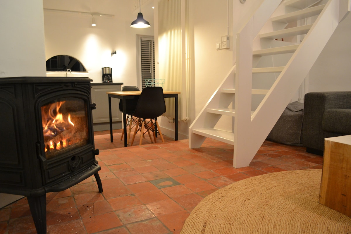 A warm living space featuring a wood-burning stove, surrounded by terracotta tiles. A dining area with a table and four chairs is seen in the background, along with a staircase leading to the upper level. Soft lighting enhances the cozy atmosphere.
