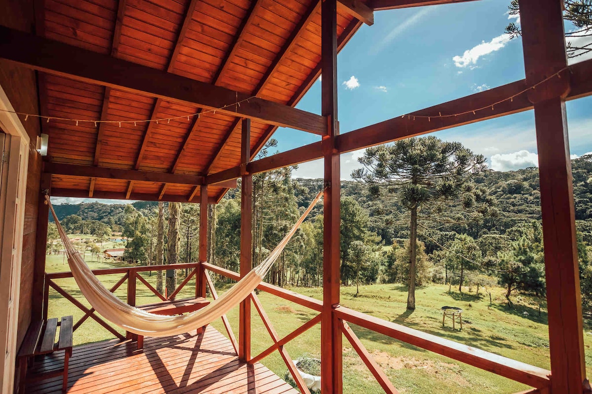 A spacious balcony features a comfortable hammock, inviting relaxation. The wooden structure captures expansive views of rolling hills and lush greenery under a clear blue sky. Sunlight streams through the open spaces, creating a serene outdoor atmosphere.