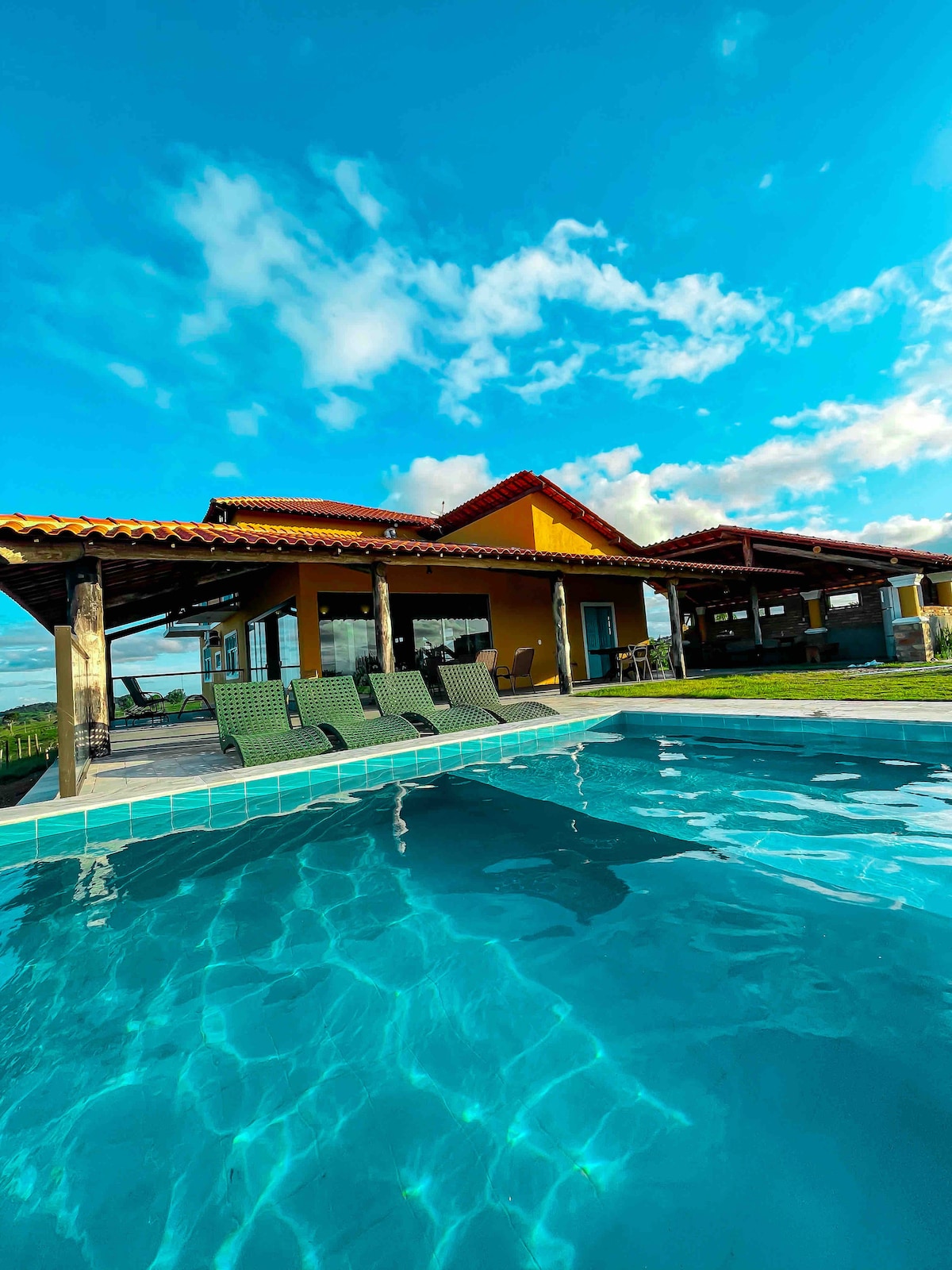 A clear blue pool reflects the sky, positioned next to a spacious outdoor area featuring a yellow house with terracotta roofs. Lounge chairs are arranged around the pool, inviting relaxation. The surrounding landscape showcases greenery under bright, partly cloudy skies.