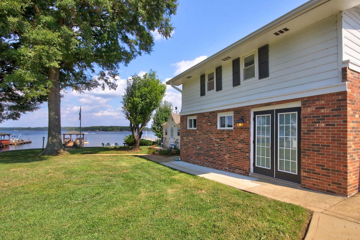 The exterior of the studio apartment is showcased, featuring a welcoming brick facade and large glass doors. A grassy area leads to the serene waters of Lake Norman, with a glimpse of the covered dock and nearby trees providing shade.