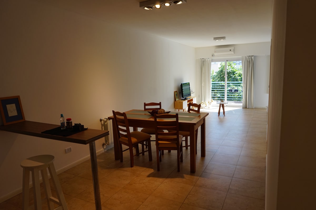 An open living space is showcased, featuring a dining table surrounded by four wooden chairs. A small side table stands nearby, and a television is mounted on the wall opposite ample windows, allowing natural light to illuminate the room. Neutral-toned tile flooring contributes to the airy atmosphere.