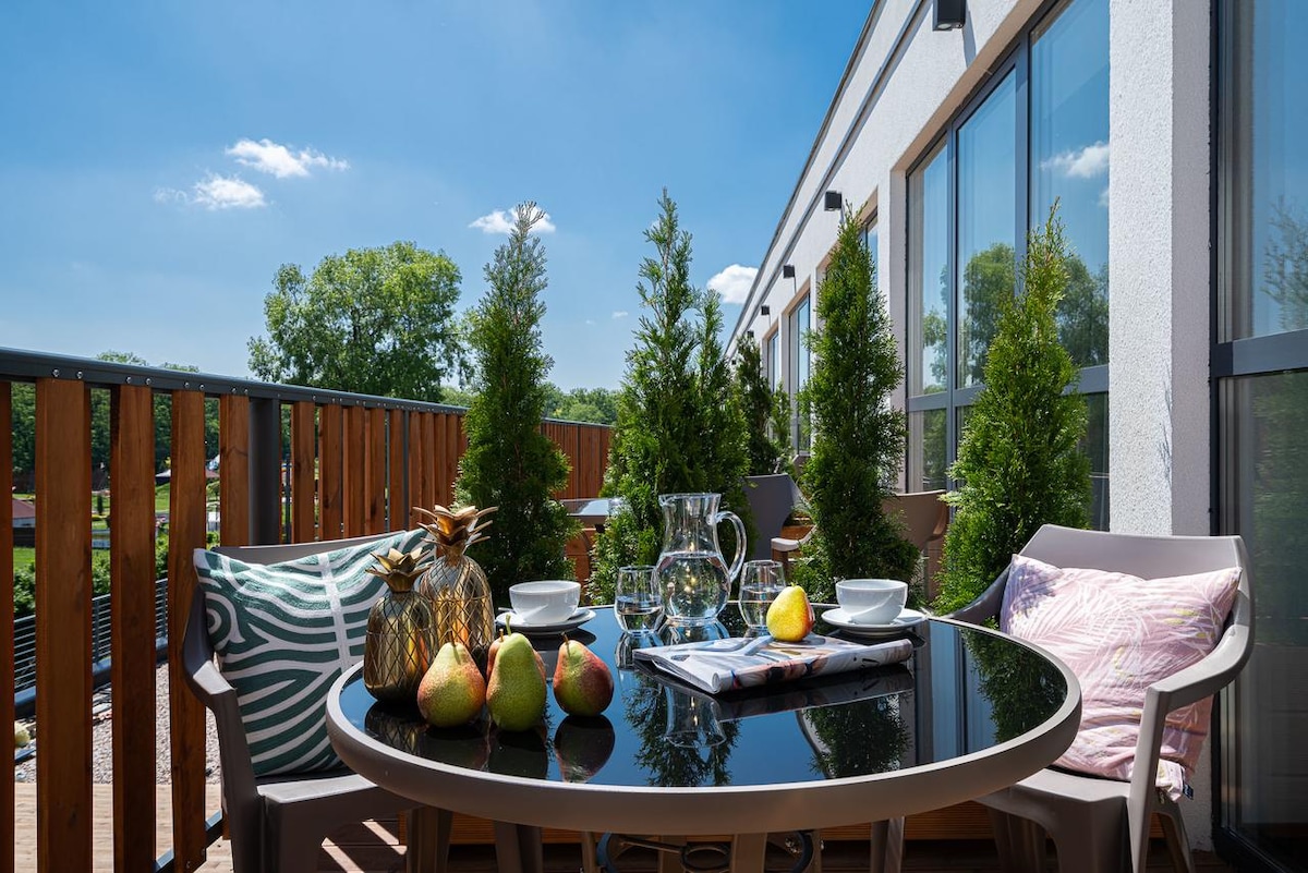 A terrace area is presented with a round black table, four white cups, a glass pitcher, and fresh pears. Two green plants flank the seating, with light fabrics on the chairs. Bright sunlight filters through the surrounding greenery, creating a serene outdoor space.