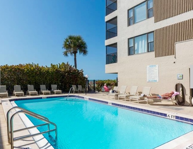 A clear view of the outdoor swimming pool is presented, surrounded by lounge chairs. Lush greenery and palm trees provide a natural backdrop. The pool's inviting blue water reflects the sunny sky above, enhancing the relaxing outdoor atmosphere.