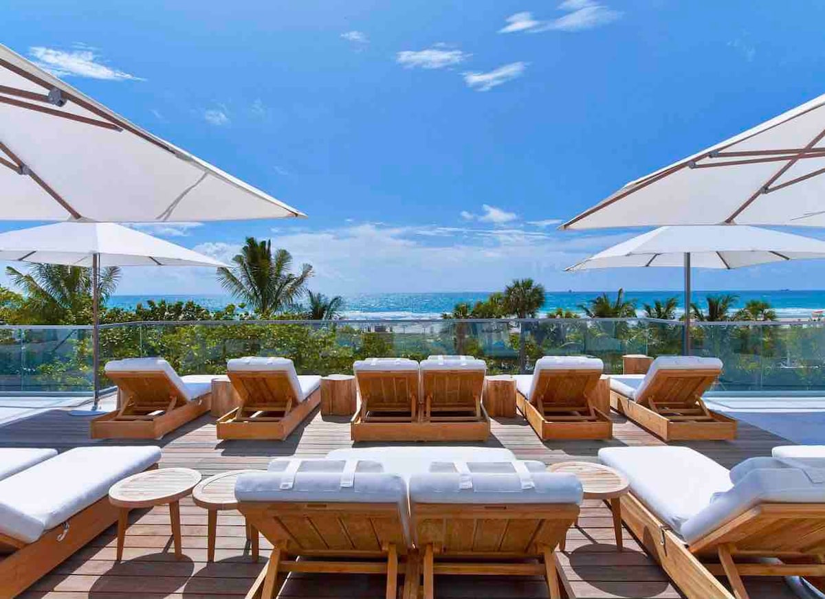 A spacious sun deck is equipped with multiple loungers and large umbrellas offering shade. The deck overlooks a vibrant ocean view, with palm trees framing the scene under a clear blue sky.
