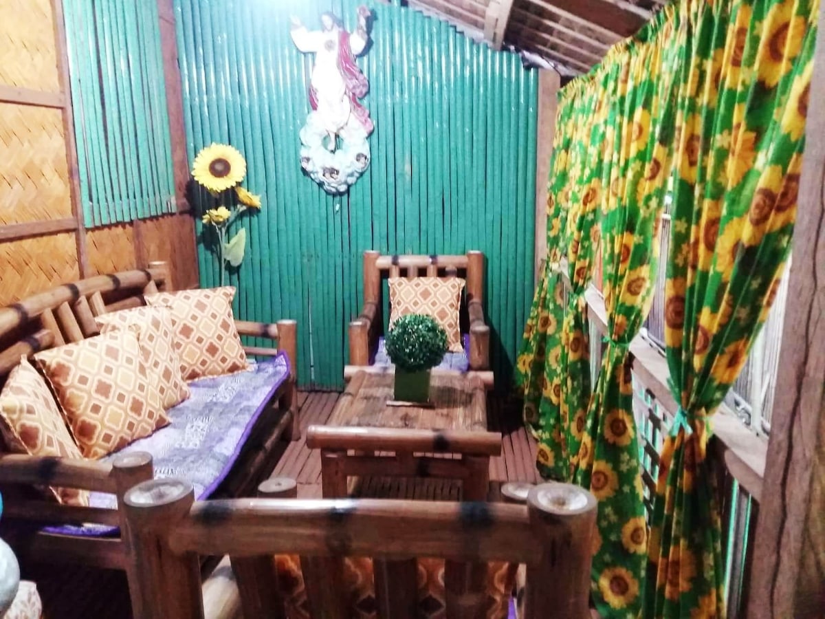 A cozy seating area is furnished with wooden chairs and a coffee table, set against a backdrop of vibrant green walls. Bright sunflower decorations are visible, along with patterned cushions on the seating, creating a warm and inviting atmosphere.