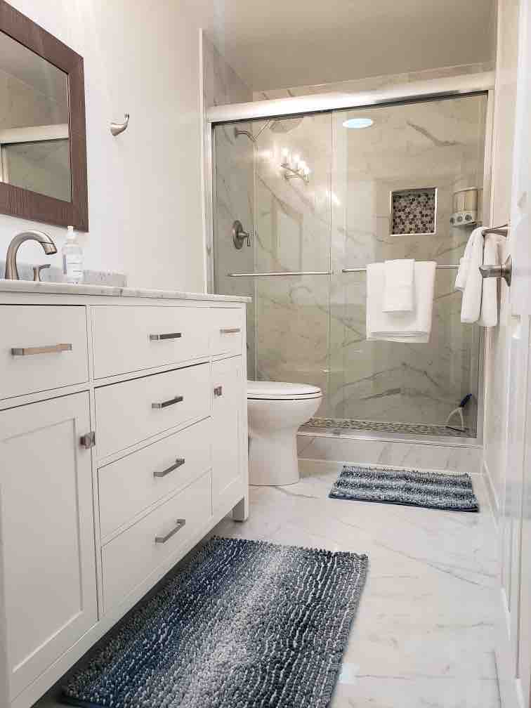 The updated bathroom features a modern vanity with multiple drawers and a stylish mirror. A glass-enclosed shower with a decorative tile inset is visible. The flooring is a sleek marble, complemented by two textured rugs placed on either side of the vanity and toilet.