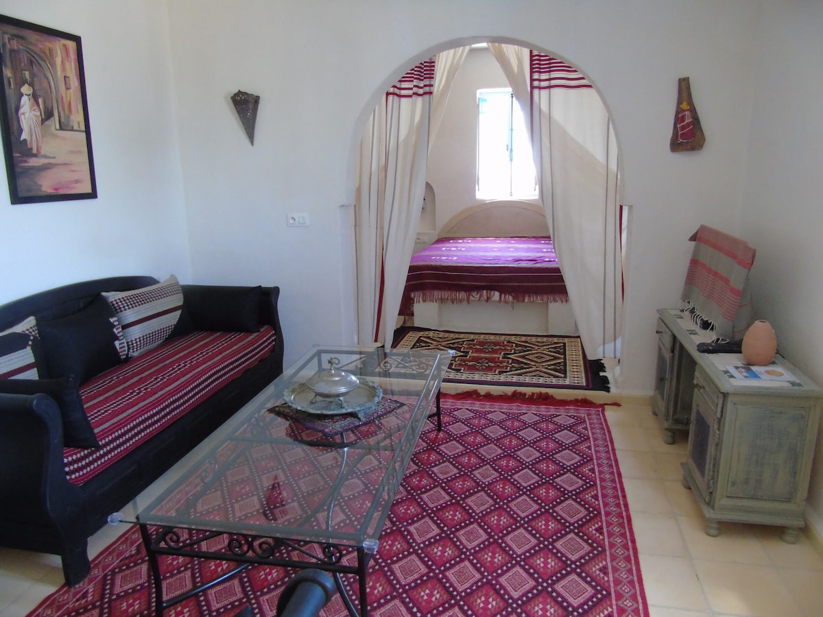 A welcoming living space features traditional decorations and a patterned rug. A glass table sits on the floor, surrounded by a black couch adorned with decorative cushions. An arched entryway leads into a bedroom, where a bed with a colorful cover can be partially seen.