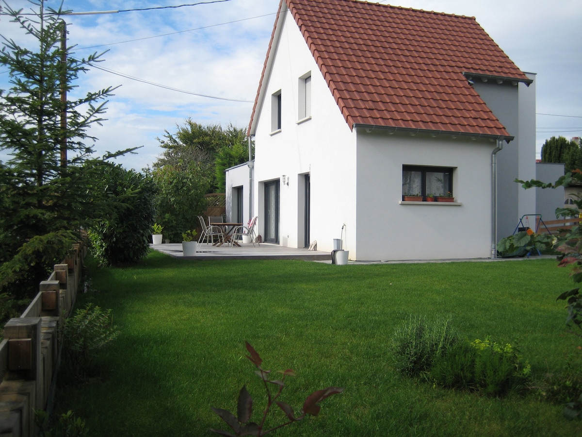 A modern two-story house is depicted, featuring a sloped red roof and white exterior walls. Adjacent to the house, a wooden terrace is visible, furnished with a dining table and chairs. The surrounding garden is green and well-maintained, adding to the inviting outdoor space.