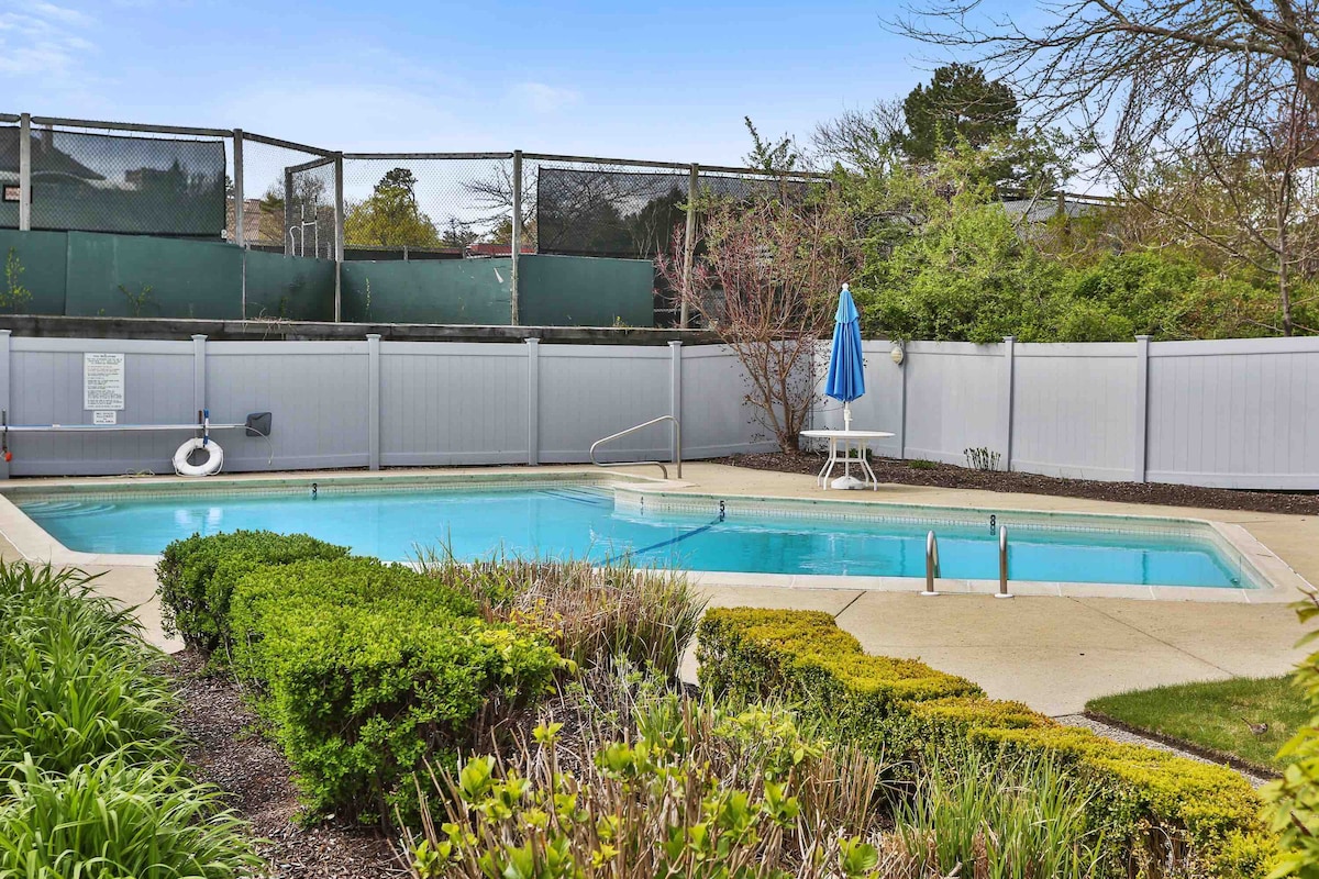 A swimming pool is bordered by neatly trimmed hedges and colorful flower beds. The pool is surrounded by a secure fence, featuring a blue umbrella on a nearby lounge chair, adding to the outdoor relaxation space.