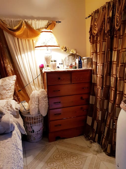 A Large Chest Of Drawers with a Lamp, near a Large window. Completed with a basket which contains extra beddings, and sleep masks.