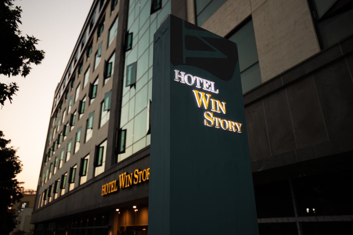 The exterior of Hotel WIN STORY is captured at twilight, showcasing a modern building with multiple floors. The illuminated hotel sign prominently displays the name, while warm light reflects from the ground-level entrance, welcoming guests as dusk sets in.