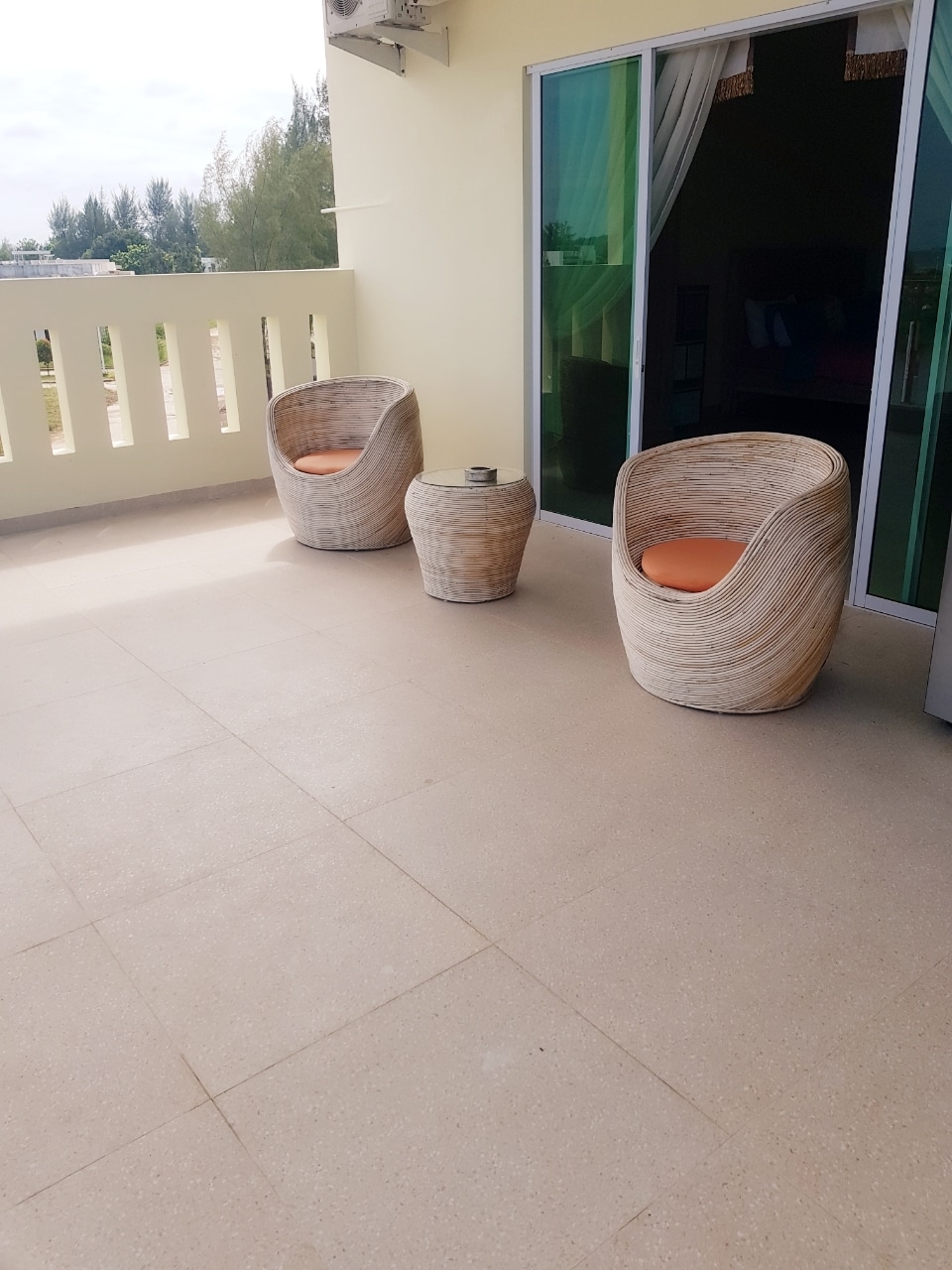 A private balcony is showcased with two curved, natural-textured chairs and a small round table. The space features smooth, beige tile flooring and an unobstructed view of the outdoors, with greenery visible in the background.