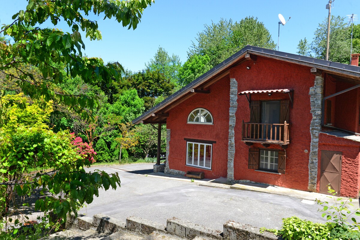 A red villa is set among lush greenery, featuring a combination of stone and wood accents. Large windows, including a prominent round one, allow natural light to brighten the interior. The parking area is spacious, enhancing accessibility to the residence.
