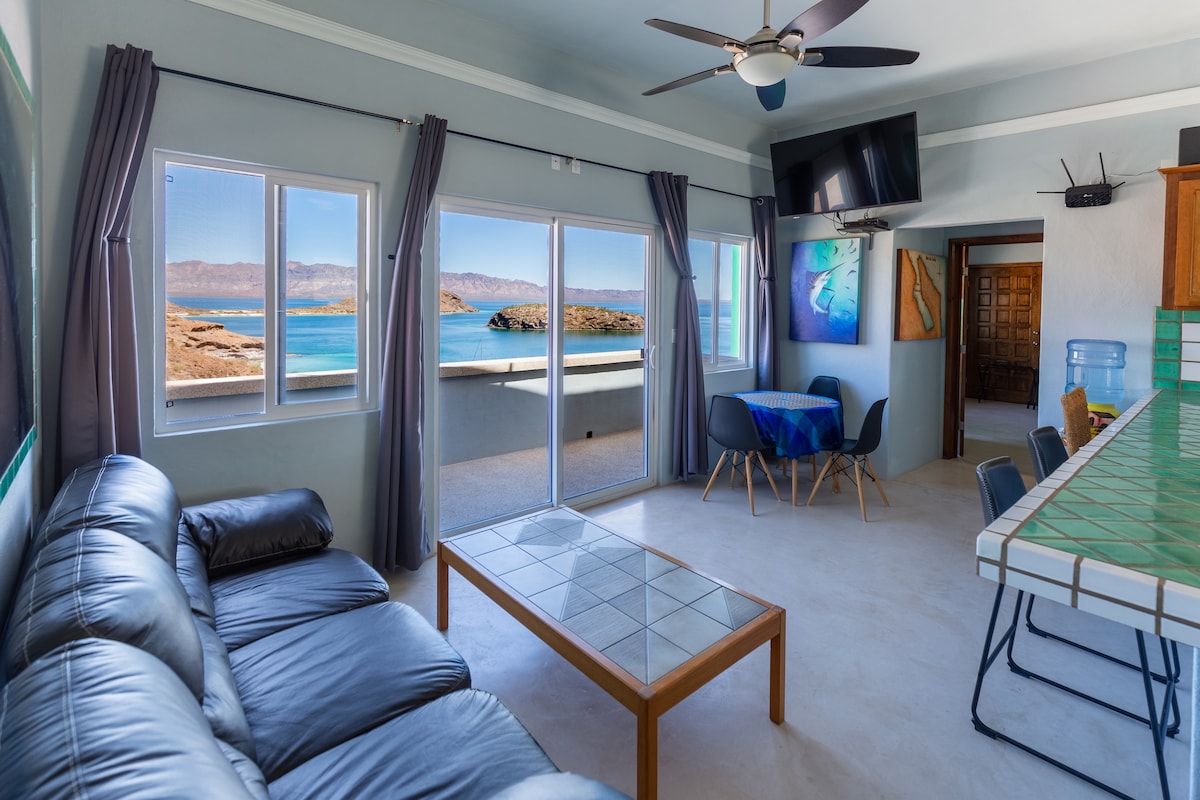 A spacious living area features a black leather sofa and a wooden coffee table. Large windows frame stunning panoramic views of the bay, with sunlight illuminating the space. A dining area with a patterned tablecloth is nearby, and a ceiling fan complements the natural light.