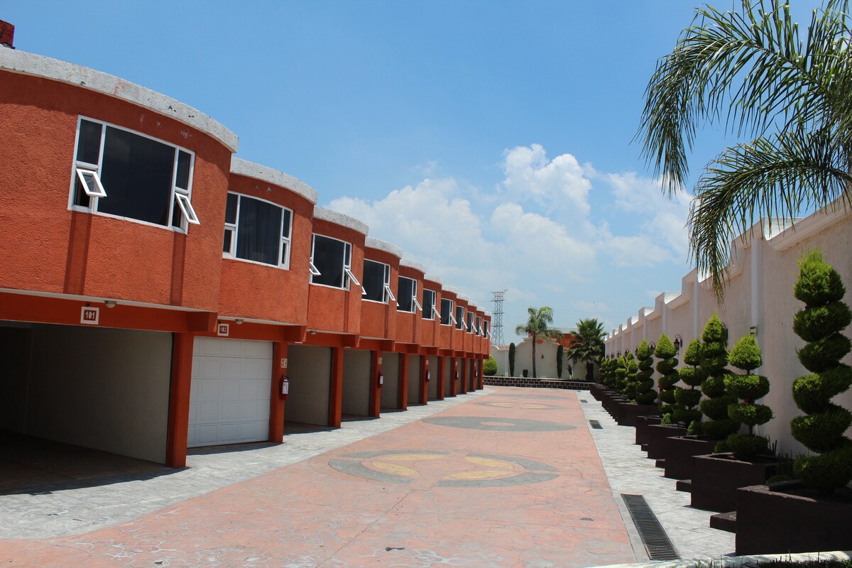 The image shows a row of modern accommodations featuring rounded facades and large windows. An open parking area runs alongside, with neatly trimmed shrubs lining the path. The sky is bright and partly cloudy, emphasizing the spaciousness of the exterior layout.