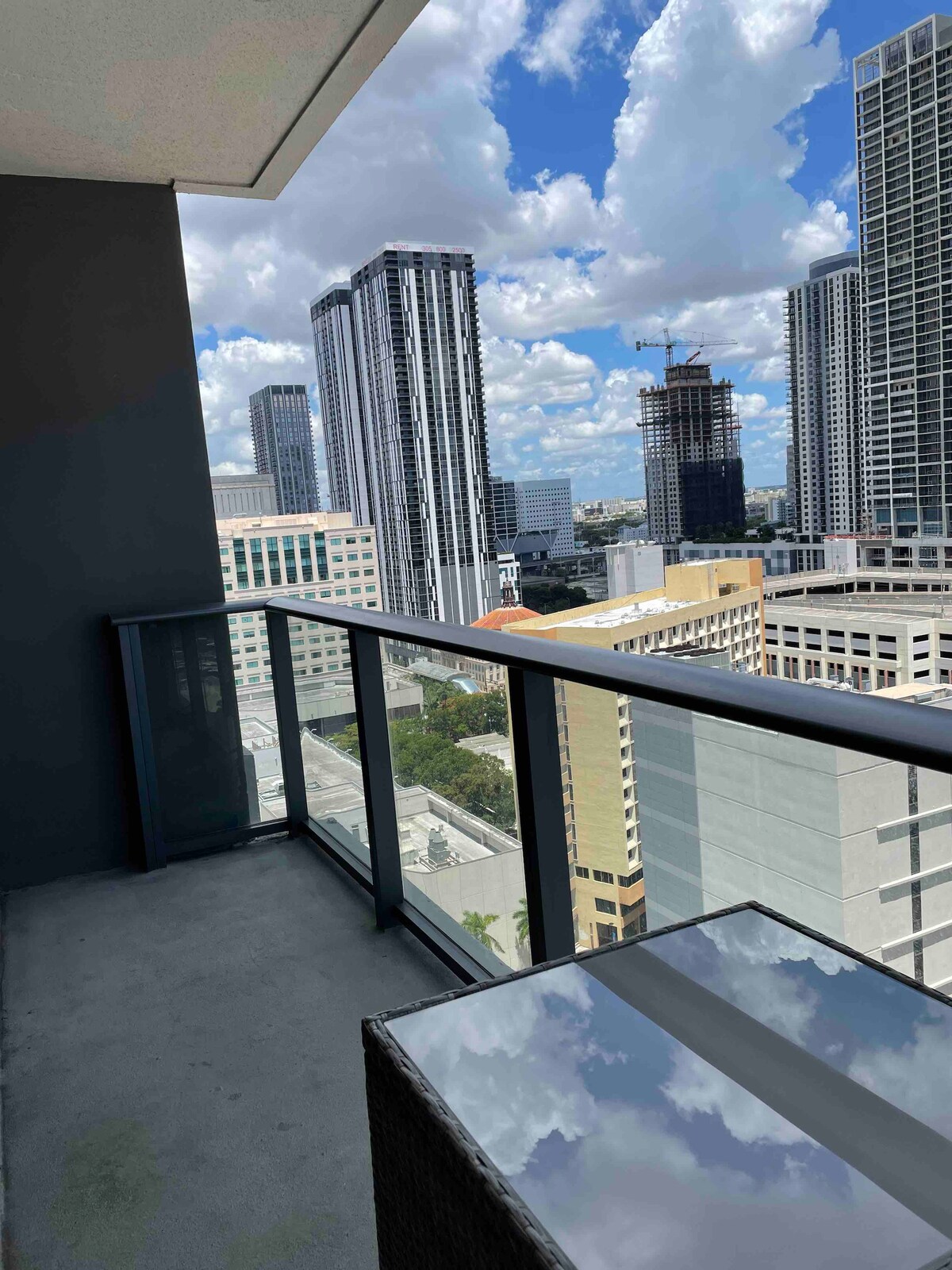 A balcony is visible, featuring a modern railing and a reflective tabletop. The view showcases a mix of tall buildings and a dynamic sky filled with clouds. The urban landscape indicates a central city location.