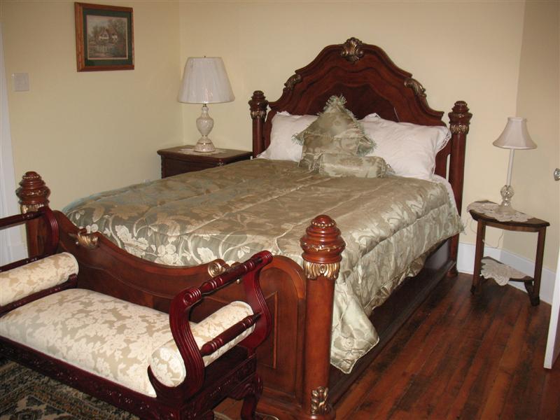 A queen size bed with ornate woodwork and a decorative comforter is centered in the room. An antique bench is positioned at the foot of the bed. A bedside table with a lamp complements the setting, along with a framed artwork on the wall.