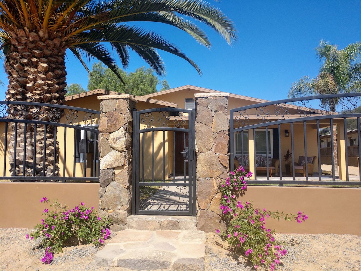 A stone pathway leads through a decorative iron gate, surrounded by vibrant pink flowers. Palm trees provide shade beside the entrance of a warm yellow house, which features large windows that invite natural light into the space.
