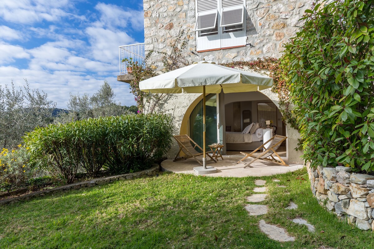 An outdoor seating area features two folding chairs beneath a large umbrella, set on a patio surrounded by greenery. A stone pathway leads from the patio, with views of the rolling landscape and olive trees, while large glass doors provide access to the room inside.