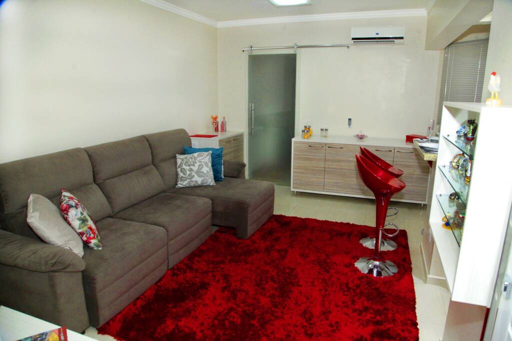 Sala ampla com visão do sofá retrátil grande, móveis com louças , taças e copos cadeiras em bancada , ar condicionado, e nicho com suvenir de vários outros países.