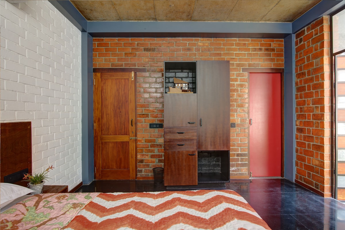 A bedroom features a cozy bed adorned with a patterned blanket, complemented by exposed brick walls. Storage options include a wooden wardrobe and a multi-drawer unit. Natural light enters through a large window, creating a warm ambiance, while contrasting door colors add to the room's character.