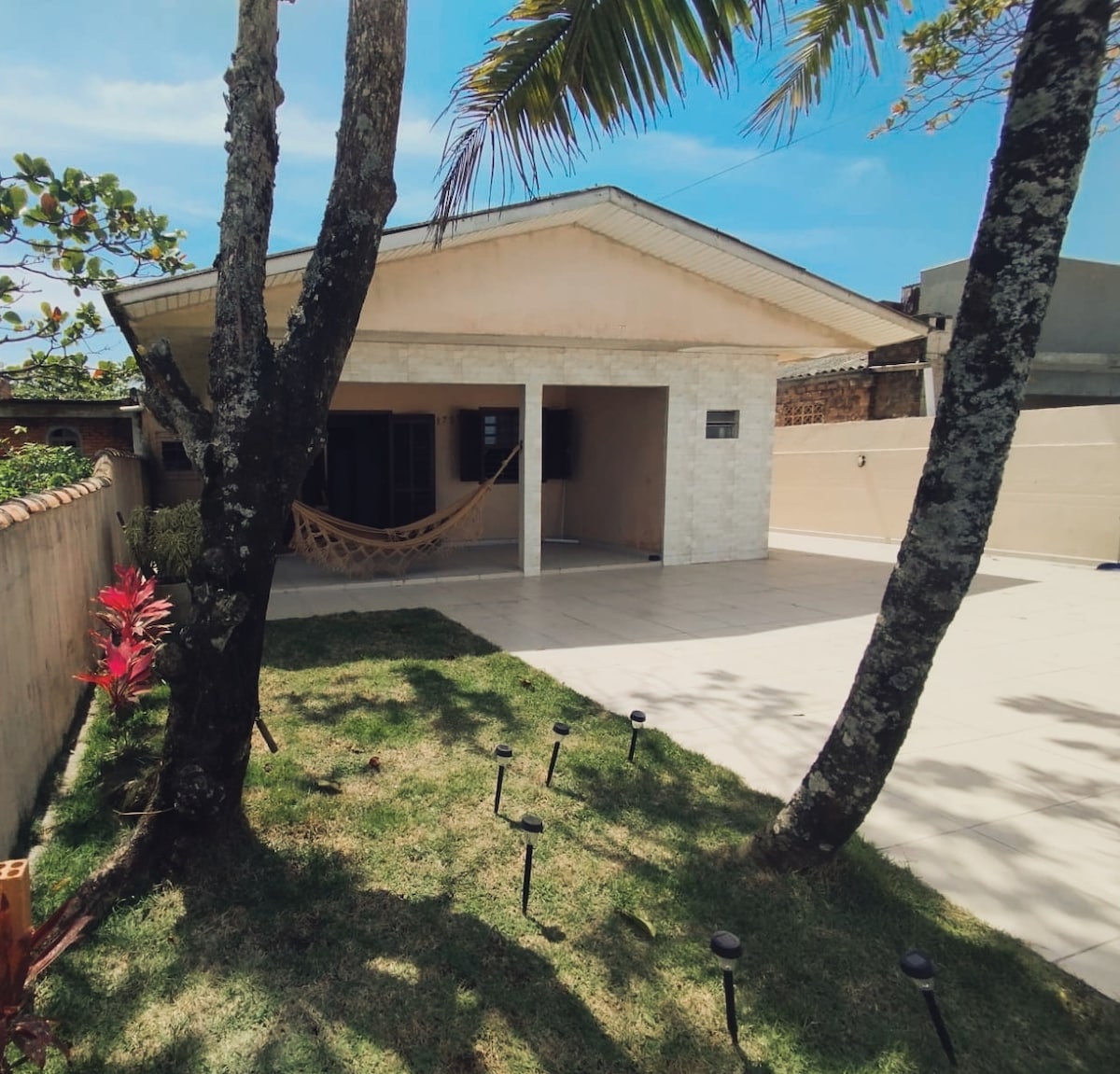 The exterior of a simple house is framed by palm trees and vibrant green grass. A covered patio area provides shaded space, while a hammock is hung for relaxation. Light-colored stone and a clear blue sky contribute to a serene atmosphere.
