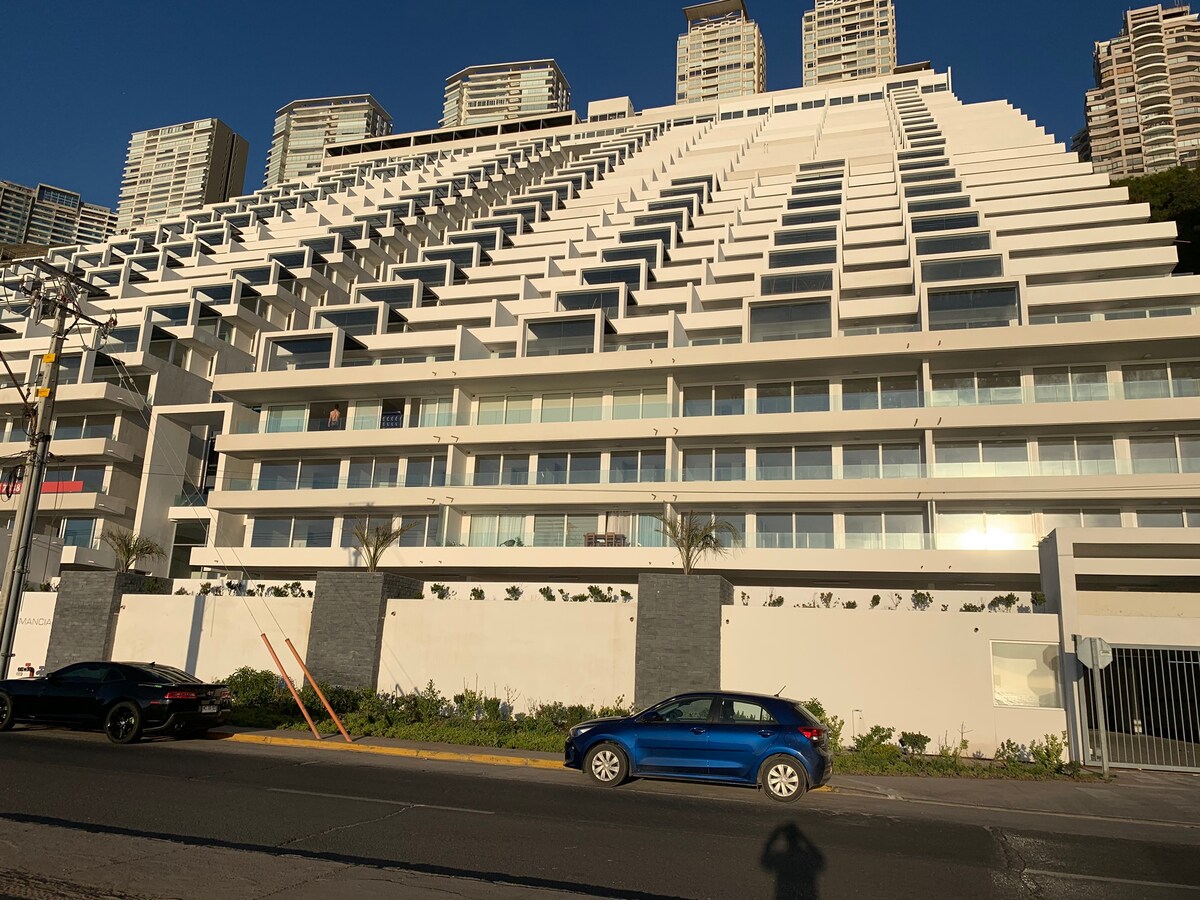 The exterior of a modern building is presented, featuring a unique stepped architectural design. Large glass windows are arranged symmetrically, allowing for natural light. The facade is complemented by greenery at the base and includes designated parking spaces in the foreground.