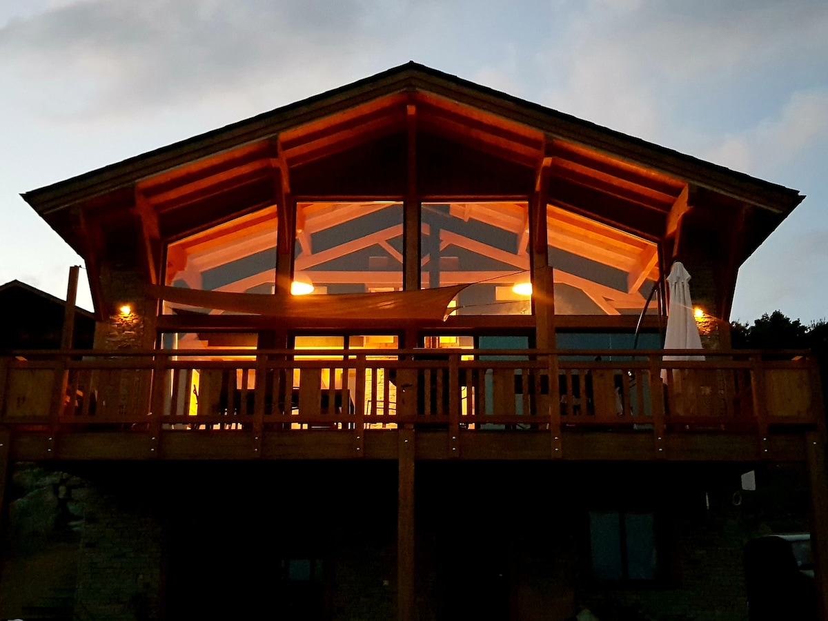The chalet's striking facade is highlighted by large glass panels, showcasing the warm glow of interior lighting. A spacious deck extends from the front, supported by wooden beams, providing an inviting entry point. The building's roof features an elegant design that enhances its contemporary style.