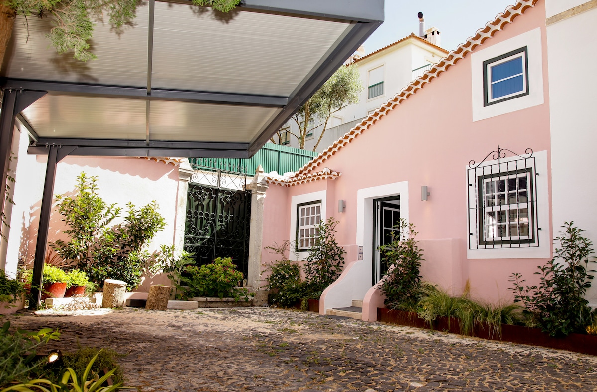 The exterior of Villa Marquês is shown, featuring a pink façade under a covered area. A stone pathway leads to the entrance, bordered by lush greenery and flower beds. Decorative elements, including wrought iron details on windows, add character to the charming architecture.