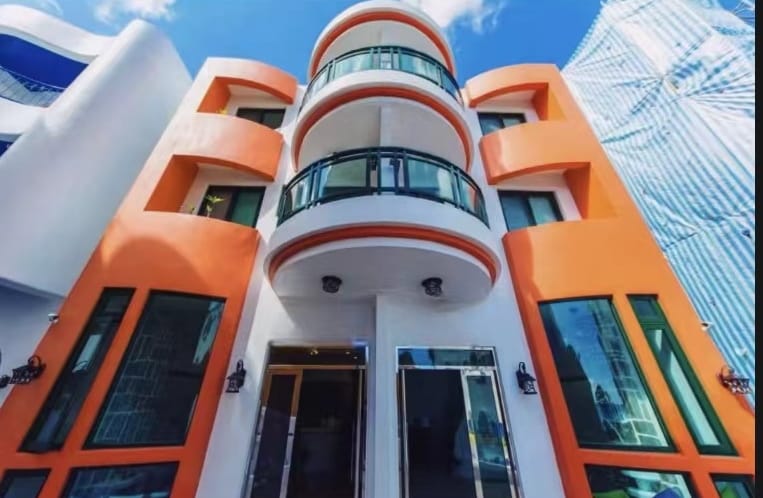 The villa features a striking facade with two floors and large windows. The vibrant orange exterior is complemented by balconies, inviting natural light into the interior. Clear skies and a hint of clouds enhance the modern architectural design.