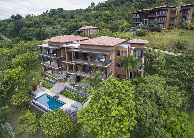 An aerial view showcases a multi-level luxury enclave surrounded by lush greenery, featuring a private pool and outdoor lounge area. The architecture blends with the natural landscape, and spacious balconies are visible on the upper floors, providing serene views of the surrounding environment.