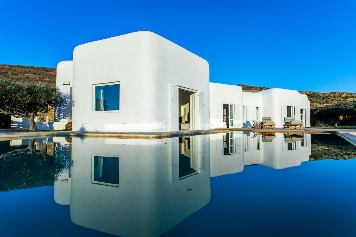 The exterior of the residence is captured under a clear blue sky, with a minimalistic white façade reflecting in the tranquil infinity pool. Large windows offer glimpses of the indoor spaces, complemented by sun loungers strategically placed around the pool area.