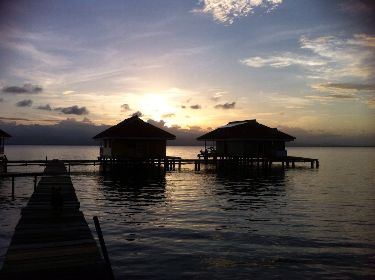 Two vacation rental villas are situated over calm waters, each featuring a thatched roof. The sun is setting behind the structures, casting a warm glow across the horizon. A wooden pathway extends towards the villas, reflective of the tranquil surroundings.