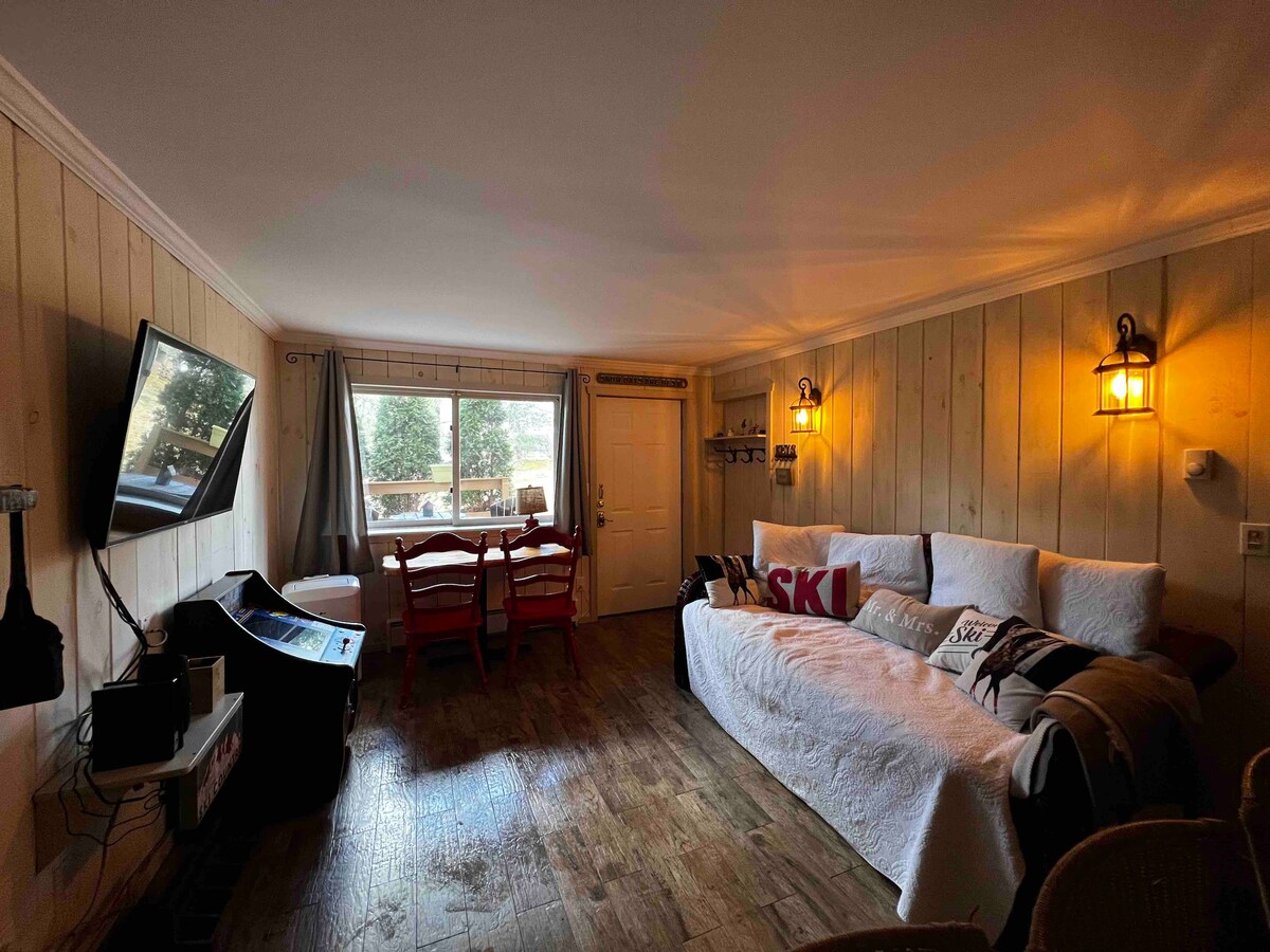 A comfortable living space is highlighted by a large window that offers natural light and views of the outdoors. A flat-screen TV is mounted on the wall, and a dining table with chairs is positioned near the entrance. Cozy blankets and decorative pillows add warmth to the seating area.