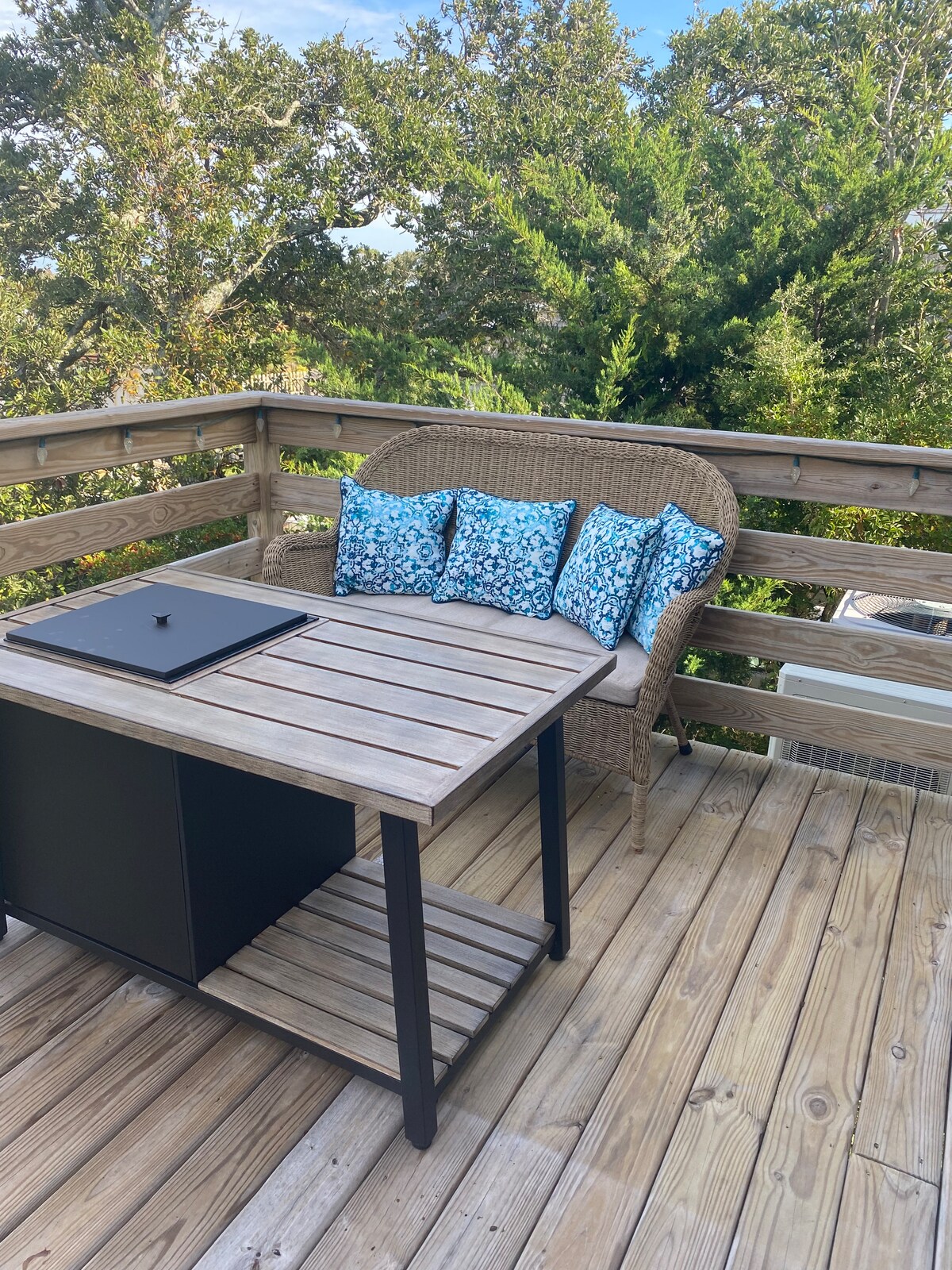 An outdoor seating area is showcased on a wooden deck, featuring a small table and a cozy wicker bench. The bench is adorned with blue and white patterned cushions. Lush greenery surrounds the space, enhancing the feeling of privacy.