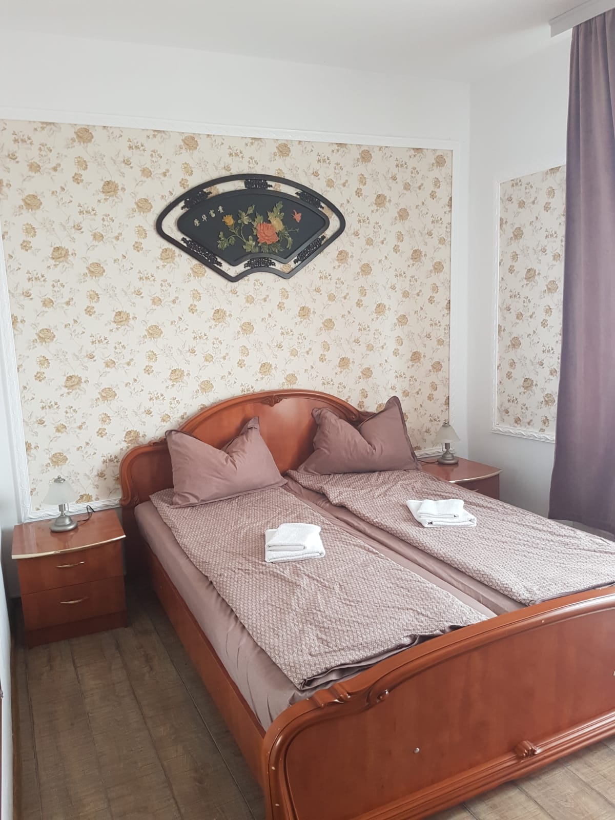 A cozy bedroom features a wooden bed with two bedding sets, complemented by decorative pillows. Nightstands flank each side, equipped with lamps. Floral wallpaper adorns the walls, while a simple decorative piece hangs above the bed. Light enters through the window, covered by a sheer curtain.