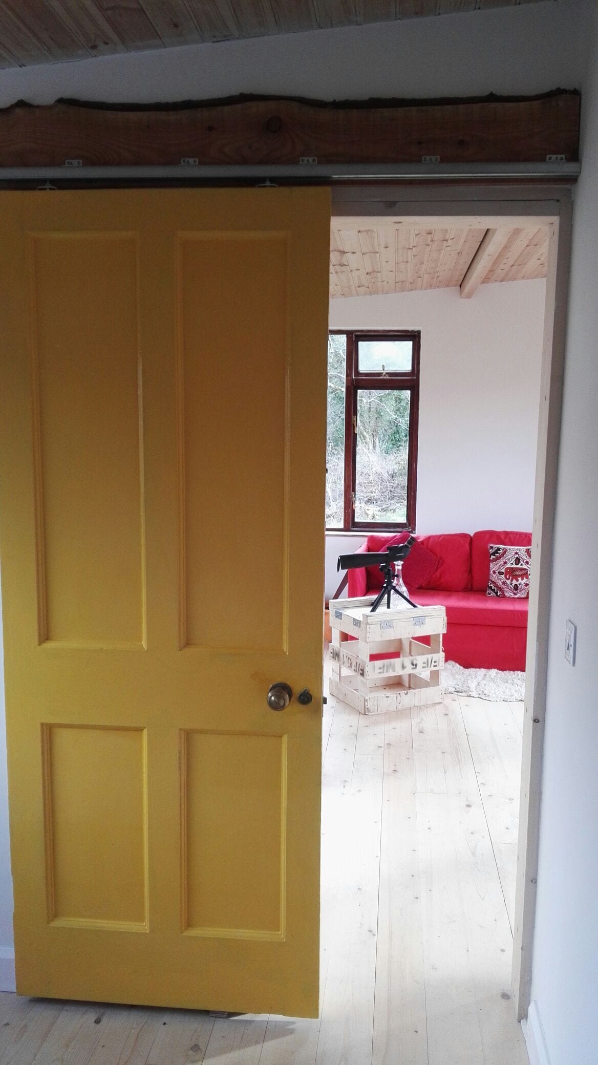 A bright yellow door is ajar, leading into a cozy living space. Inside, a vibrant red sofa is visible, accompanied by a transparent box table. Natural light enters through a nearby window, illuminating the wooden flooring and soft textures of the interior.