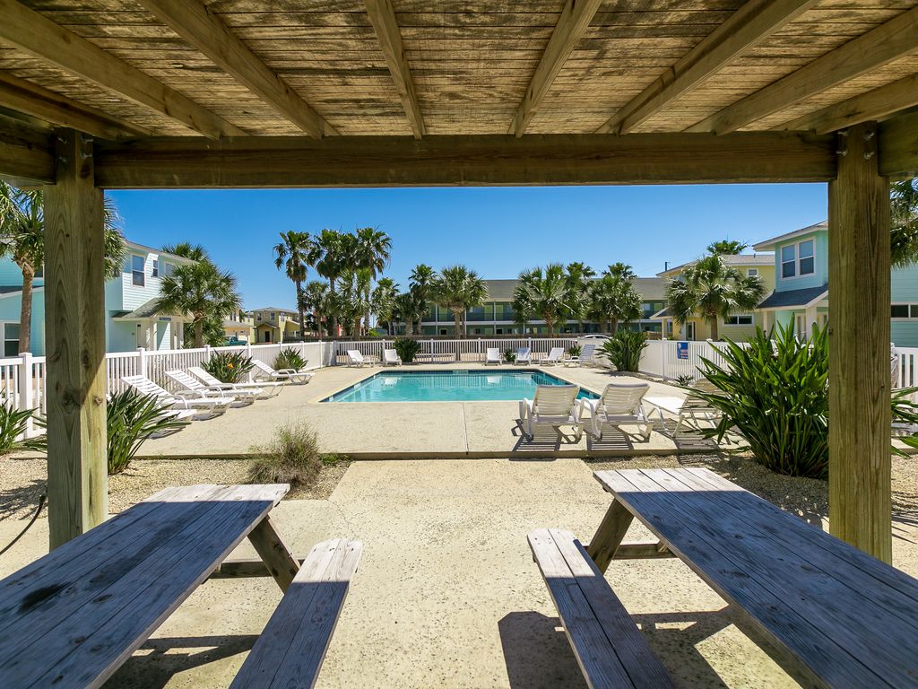 A shaded outdoor space features a picnic table overlooking a well-maintained pool area. Several sun loungers are arranged near the pool, surrounded by palm trees and landscaped greenery. The area is designed for relaxation and enjoyment in a coastal environment.