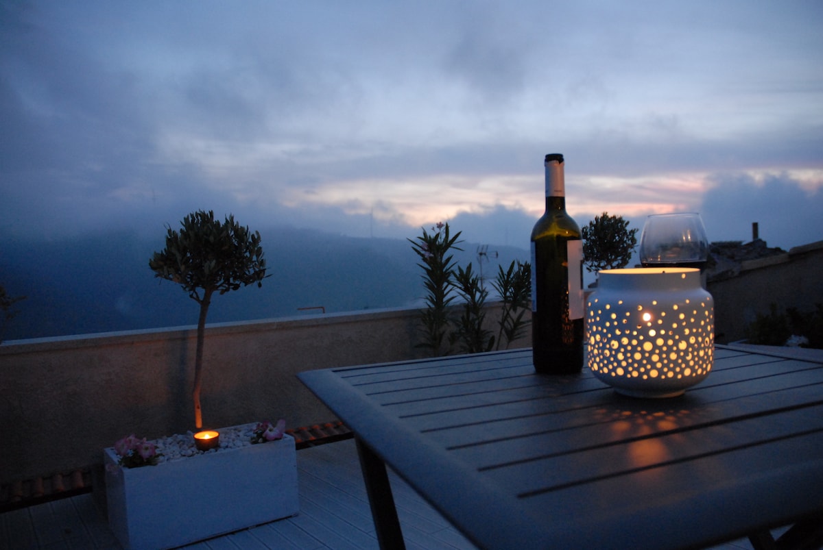 An outdoor terrace is displayed with a table set for relaxation. A bottle of wine and a glass rest on the table beside a glowing lantern. Greenery is visible in the background, while the sky showcases soft hues as the sun sets.