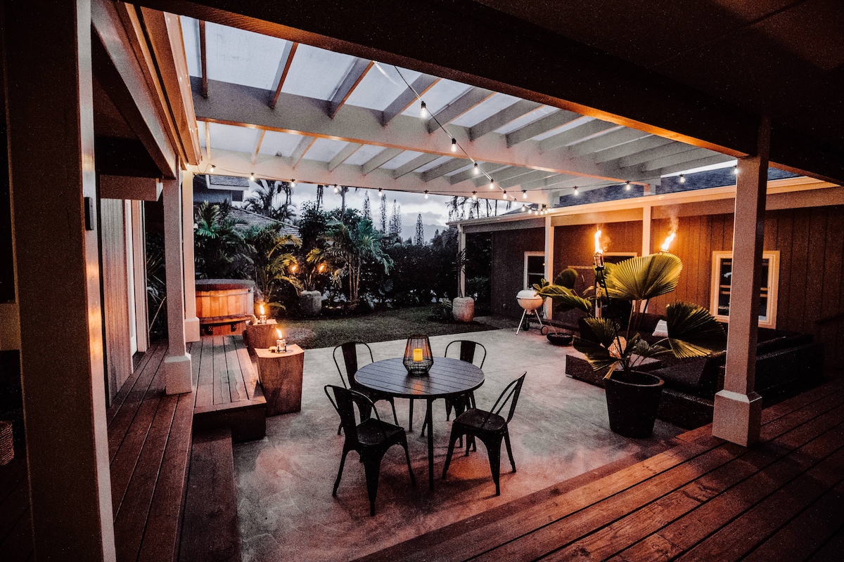 An inviting outdoor space is highlighted by a dining table surrounded by black chairs, under soft lighting from overhead fixtures. A cedar hot tub is placed in the corner, complemented by tropical plants and a cozy seating area, perfect for evening relaxation.