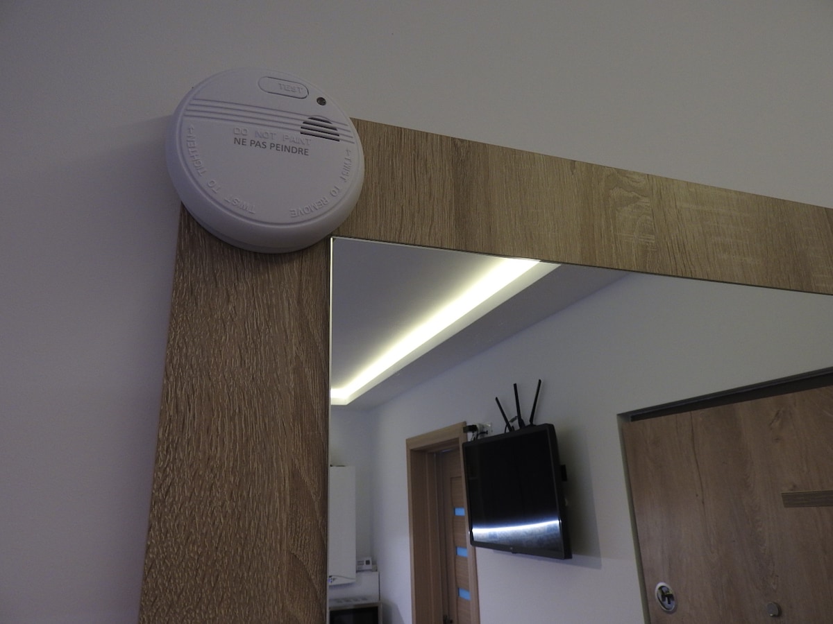 A smoke detector is mounted on the wall above a modern wooden mirror. The mirror reflects partial views of the room's interior, with a flat-screen TV visible on the opposite wall. Soft lighting is installed above the mirror, enhancing the space's illumination.
