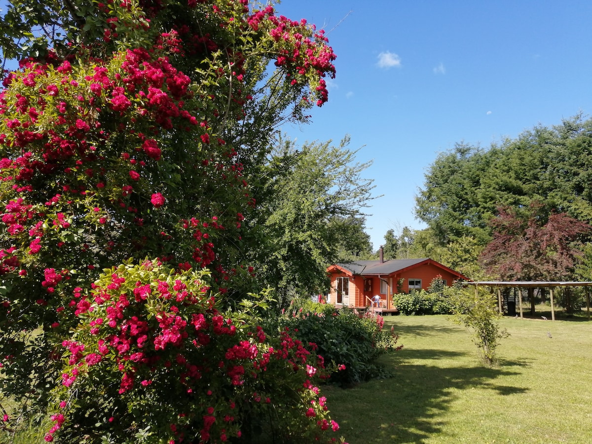 A wooden cabin is set against a backdrop of lush greenery, surrounded by flowering shrubs in vibrant pink. The expansive lawn is dotted with trees, creating a serene atmosphere. Clear blue skies are visible above, enhancing the peaceful outdoor setting.