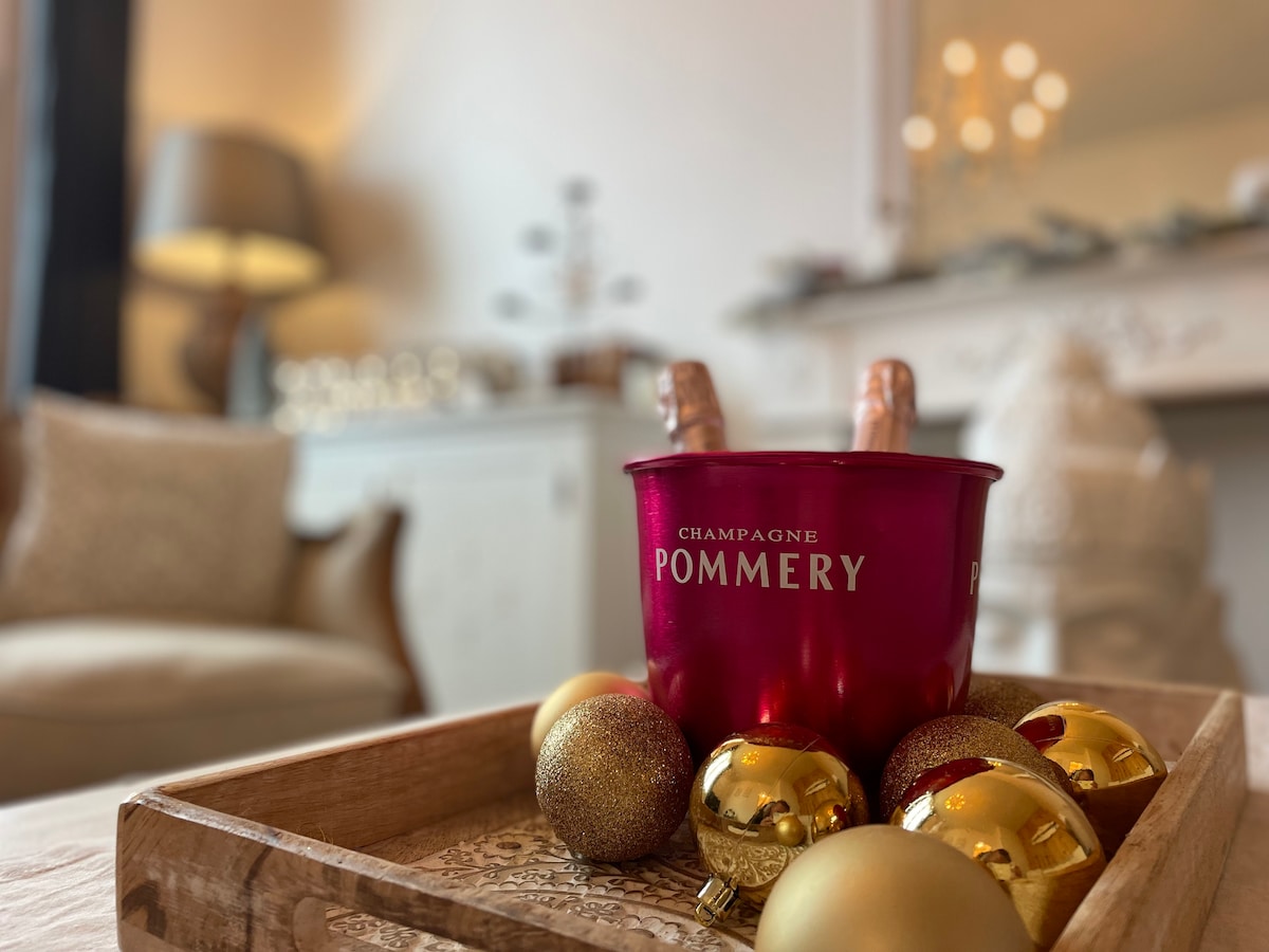 A red champagne bucket holds chilled bottles, set on a wooden tray adorned with decorative golden ornaments. Soft lighting from a nearby lamp creates a cozy ambiance in the background, highlighting a tasteful living space with neutral tones.